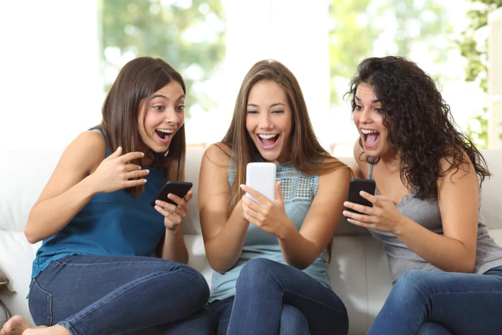 Three young women laugh while looking at social media on their phones. 