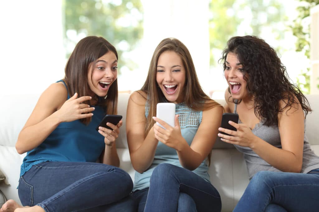 Three young women laugh while looking at social media on their phones.